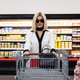 A blonde woman wearing dramatic sunglasses and a black and white blouse pushes a supermarket shopping cart in Jakob's Wife.